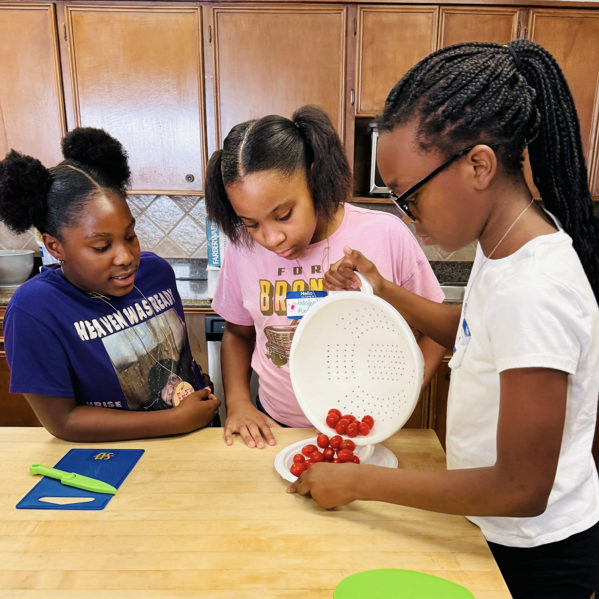 Three girls pouring cherry tomatoes out of a colander onto a plate.